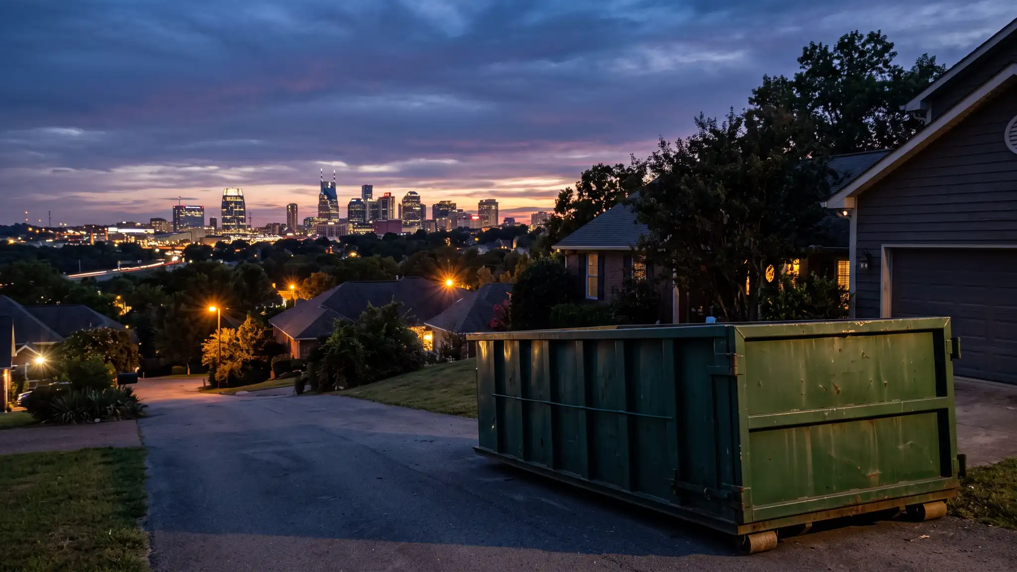 A dramatic dusk view of the Nashville skyline with warm lights over rooftops and a dark green dumpster in a suburban driveway in the foreground